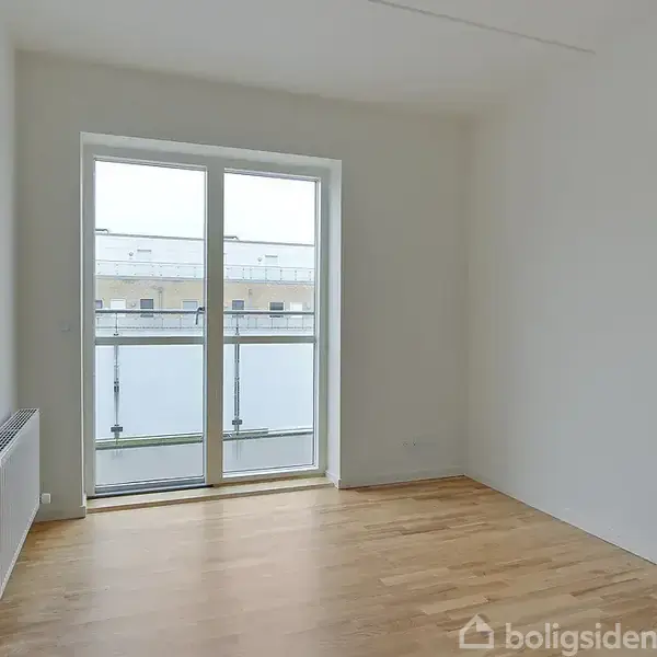 Et tomt værelse med trægulv og en stor glasdør, der fører ud til en altan. Væggene er hvide, og der er en radiator på venstre side.