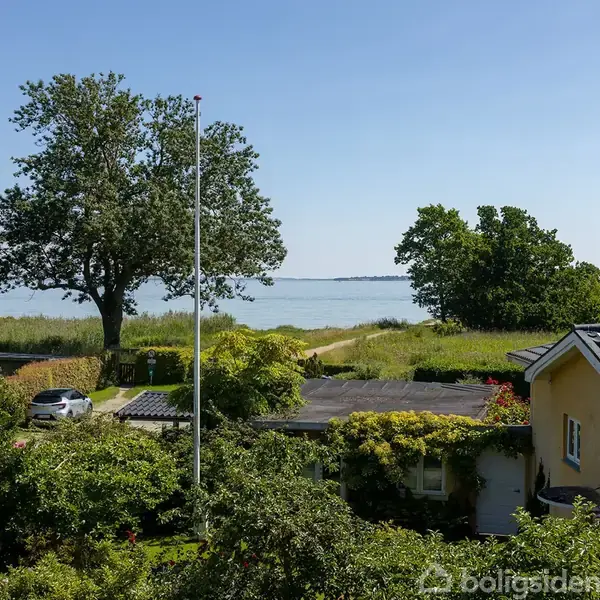 Gul villa med solpaneler på taget står i en frodig have. I baggrunden ses en vej, der fører til kysten med udsigt over havet og omkringliggende træer.