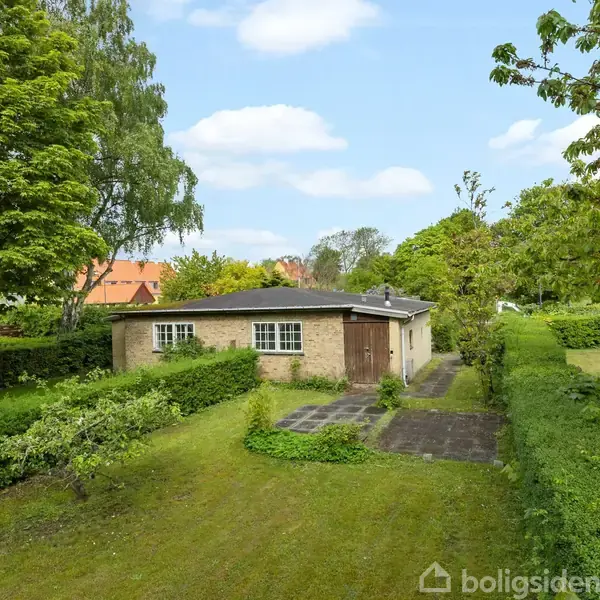 Et lille hus med fladt tag ligger i en frodig have omkranset af buske og træer. En sti fører til huset gennem den velholdte græsplæne.