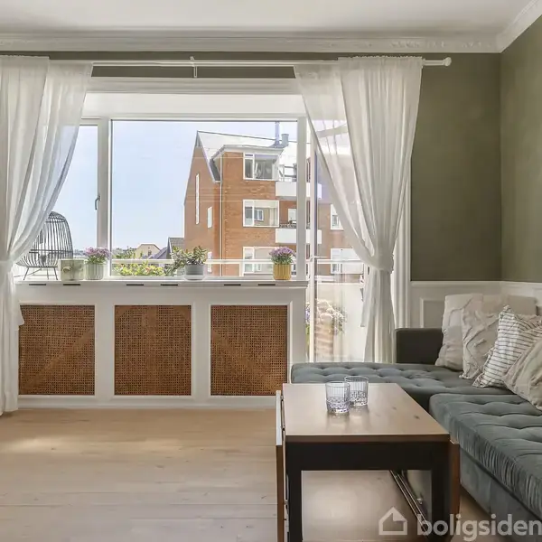 Stue med grøn sofa dekoreret med puder står ved siden af et træbord. Store vinduer med hvide gardiner slipper lys ind, og udenfor ses en bygning. Ingen tekst er vist.