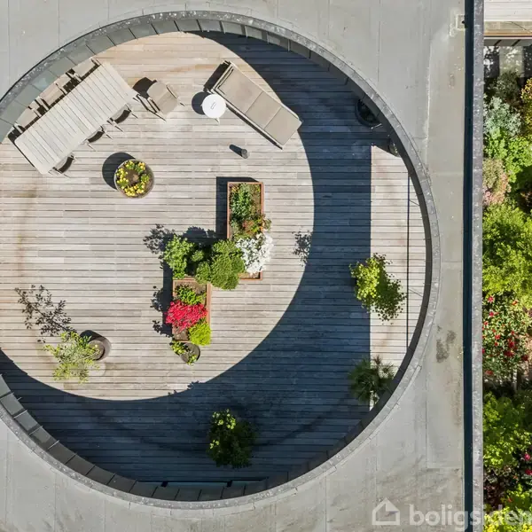 Rund terrasse med træmøbler og planter, set fra oven. Terrasse omgivet af kant af bygningen. Til venstre ses en have med forskellige buske og træer.