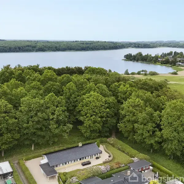 Et hus med mørkt tag ligger omgivet af frodig skov. I baggrunden ses en stor sø med en strandbred og et boligområde langs vandkanten.