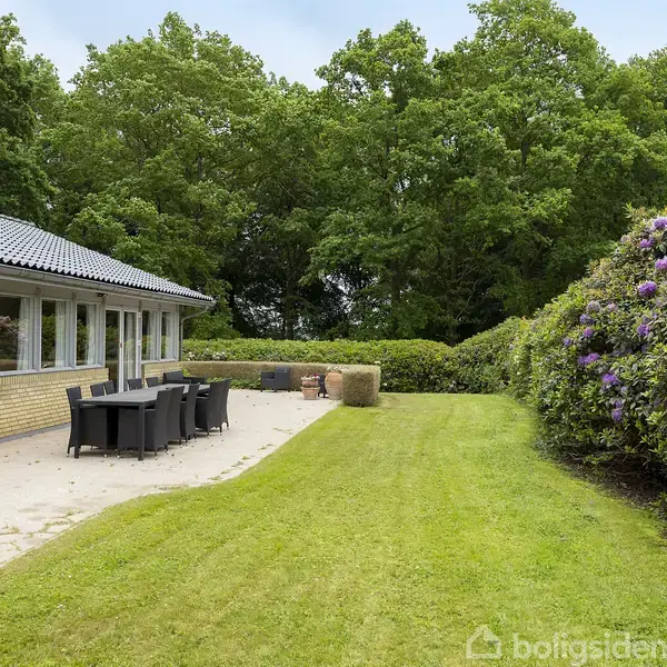 En terrasse med sort møbelsæt står ved siden af et hus med store vinduer. Omgivet af en velplejet græsplæne og blomstrende buske, danner skov baggrunden.