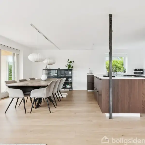 Spisebord med otte stole står i spisestue, omringet af store vinduer og moderne køkken en suite. Et sort skab og planter skaber en minimalistisk indretning i den lyse atmosfære.