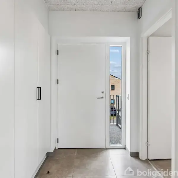 Hvid hoveddør omgivet af skabe åbner mod en lys entré med fliser og synligt udsyn til udendørs. To døre fører til nærliggende rum.