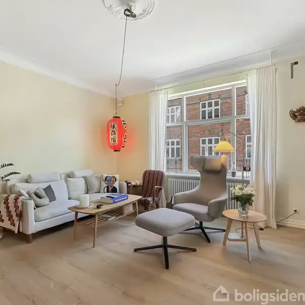 Stue med sofa, lænestol og sofabord, indrettet med en rød papirlampe. Vindue med udsigt til murstensbygning. Bogreol på højre side, maleri og plante på venstre side.