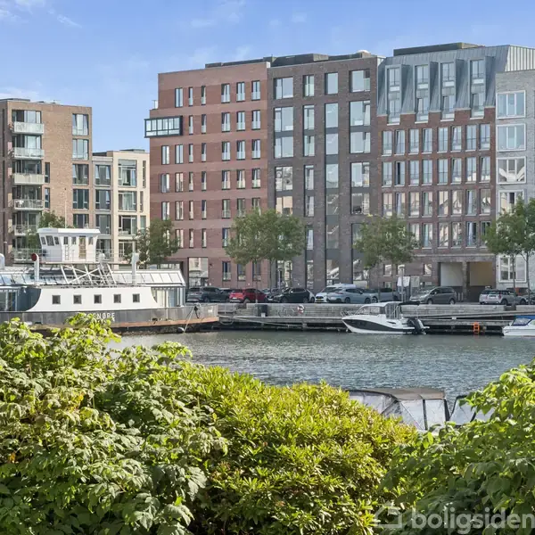 En båd ligger stille i en kanal omgivet af moderne, flerfarvede bygninger. Foran er der frodig grøn beplantning. Biltrafik og både ses på vandet.