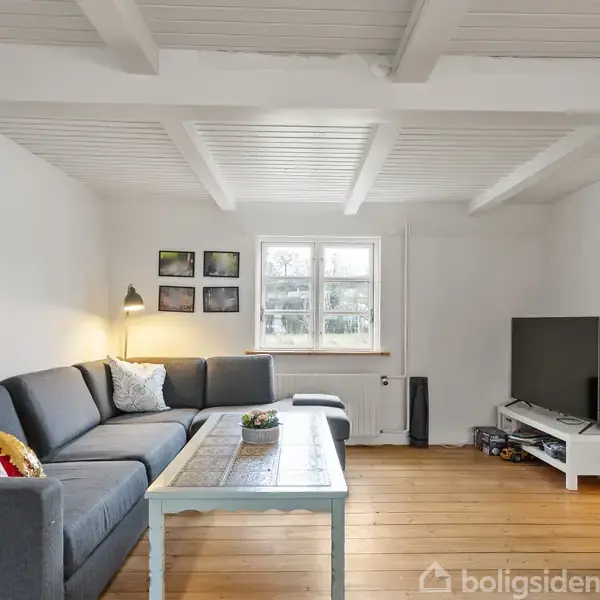 Grå sofa står i hjørne, flankeret af et lavt bord i hvidmalet stue med synlige bjælker. TV står til højre på mediemøbel, ved siden af en døråbning. Vindue i midten.