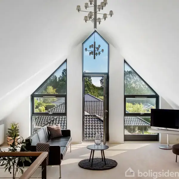 Loftstue med skråt loft og stort trekantet vindue fyldt med sollys. Sofa og tv i moderne stil står ved vinduet. Træer og tagrygge ses udenfor.