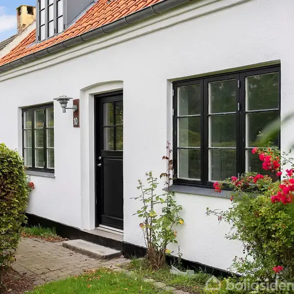 Hus med hvid facade og sort dør. Røde blomster vokser ved siden af indgangen. Området er omgivet af grønne buske og græsplæne under en blå himmel med få skyer.