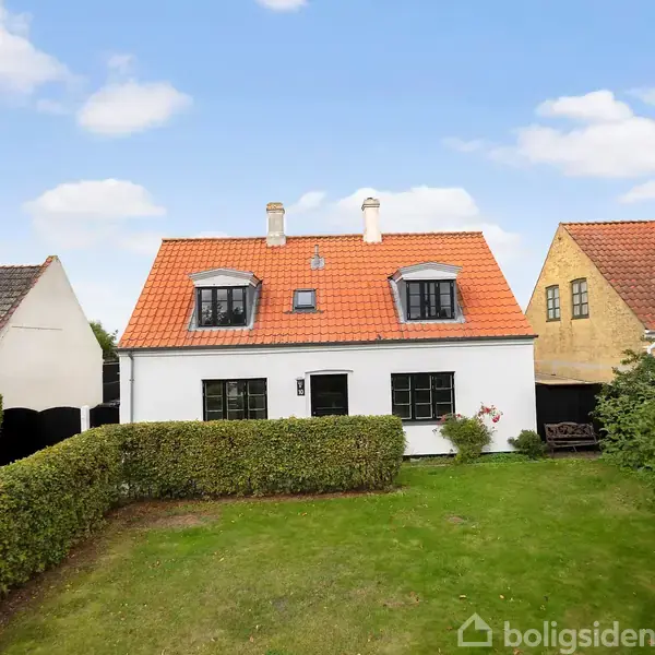 Hus med hvid facade og rødt tag står i en velholdt have med grøn græsplæne. Nabohuse ses på begge sider under en blå himmel med spredte skyer.
