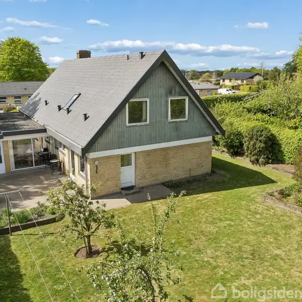 Et trekantet taghus står midt i en grøn have. En terrasse er forbundet til huset. Omgivet af hække og træer, ligger huset i et roligt boligområde.