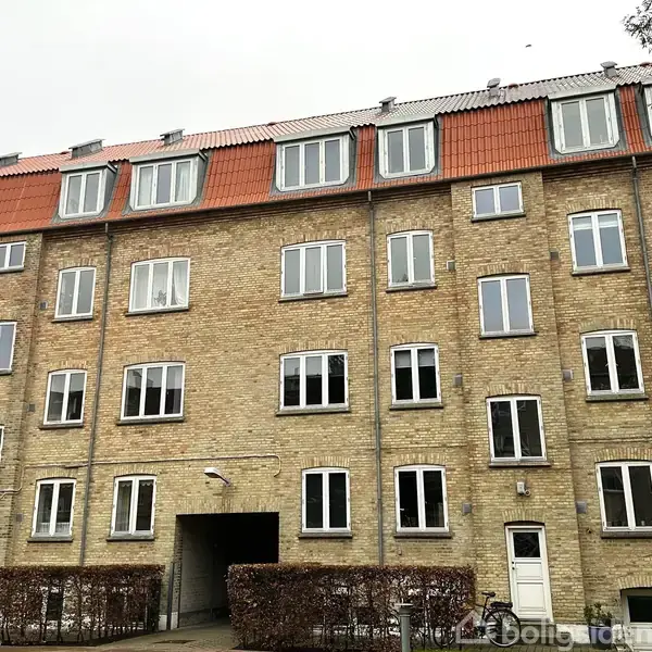 En stor murstensbygning står med flere etager, alle med hvide vinduer og en rødt tag, beliggende i et beboelsesområde. Hække omkranser bygningens base og en cykel er placeret nær en indgang.