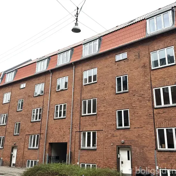 Rød murstensbygning med flere vinduer strækker sig langs en stille gade. Lygtepæl hænger over vejen. Boligen har flere etager med rødt tag. Busk i forgrunden.