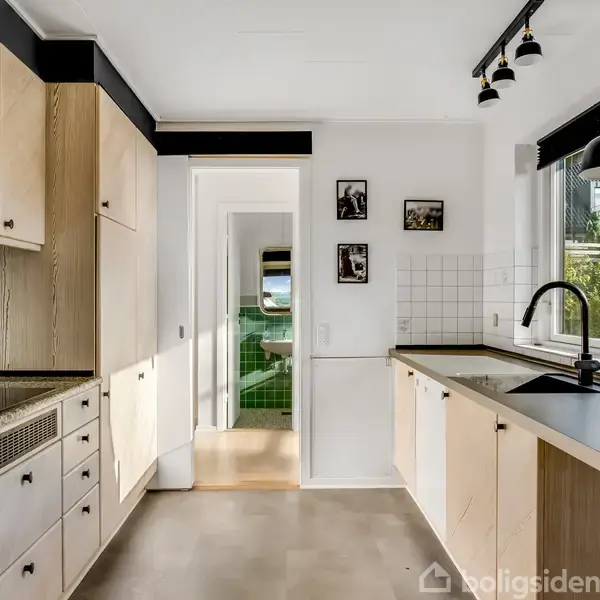 Køkken med lyse træskabe og sorte bordplader. En sort vandhane stikker ud over en vask under et stort vindue. Klinker og billeder på væggen tilføjer detaljer.