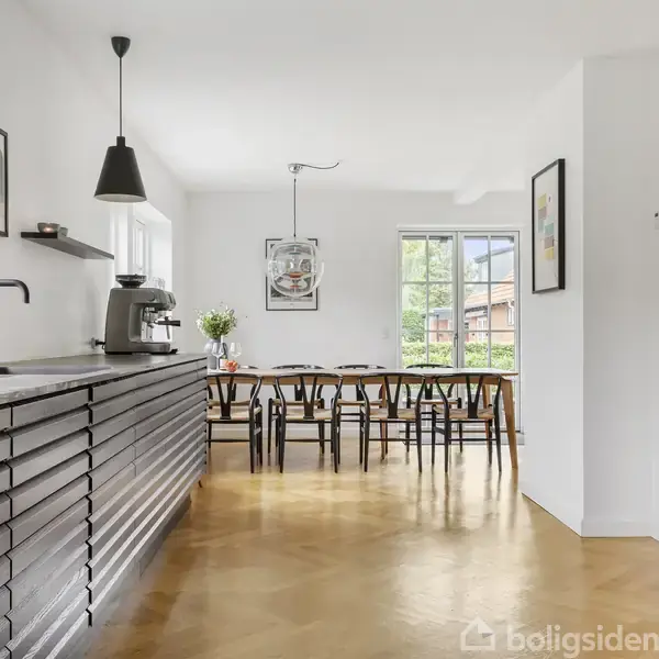 Køkken med sort skabsmodul, vandhane og espressomaskine på en træbordplade. I baggrunden et spisebord med sorte stole, omgivet af hvide vægge og store vinduer, der slipper lys ind.