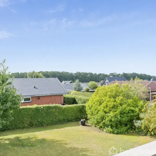 Husene med skrå tage står i et boligområde med velplejede haver. Et stort træ og buske omgiver en grøn græsplæne, mens en blå himmel strækker sig over den landlige udsigt.