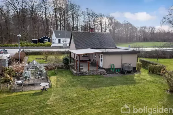 Huset med en overdækket terrasse står i en velplejet have med et drivhus og borde. Baggrunden består af en vej, træer og marker under en blå himmel.