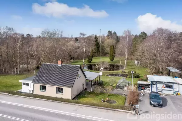 Et enkelt hus med gråt tag står ved en landevej. Ved siden af huset er der en have med et drivhus og en dam. Skov fylder baggrunden.