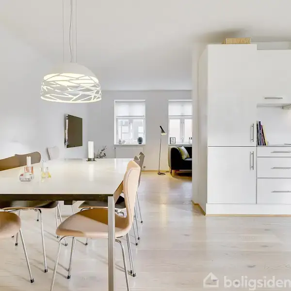 Spisebord med seks træstole står i en lys spisestue. En moderne lampe hænger over bordet. Baggrunden viser et åbent køkken med hvide skabe og en ovn.