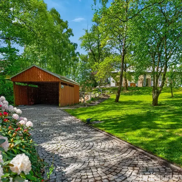Bænk står ved husets mur, under et blomstrende træ. Fliser dækker jorden og en stor blomsterpotte med planter står ved siden af bænken. Et vindue og en buet passage ses i baggrunden.
