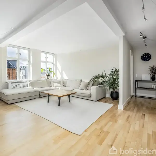 Stor, lys stue med hvid sofa omkring et træbord. Store vinduer lader naturligt lys komme ind. En plante står til højre, og en vægdekoration samt ur er synlige.
