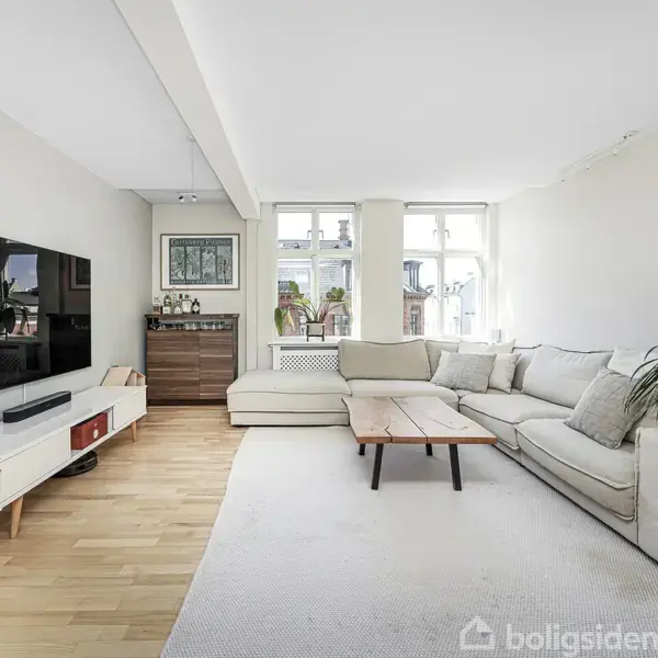 En lys stue med en stor, beige sofa står i hjørnet. Sollys strømmer ind gennem vinduerne. En grøn plante og et lille sofabord supplerer det hyggelige rum.