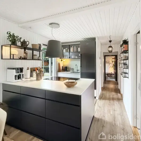 Køkken med sort køkkenø og hvide vægge, hvor planter og køkkenredskaber står på hylder. Et spisebord med stole er synligt, og en gang leder til andre værelser.