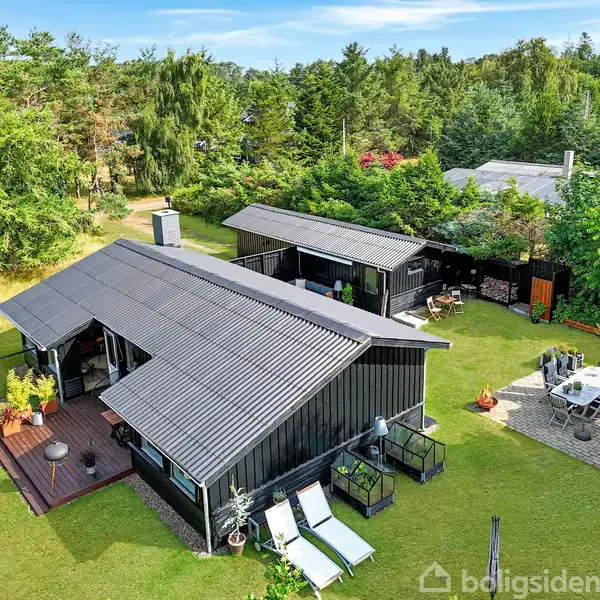 Et sort træhus med terrasse ligger i en frodig have. Ved siden af findes et område med havebord og parasol. Skovområder omgiver ejendommen i baggrunden.