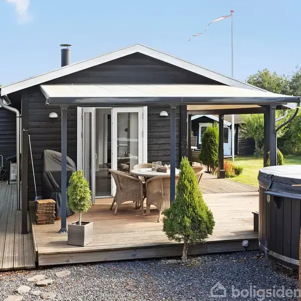 Et sortmalet træhus med overdækket terrasse, hvor et bord og stole står. Omgivet af grønne planter i en have på en solrig dag.
