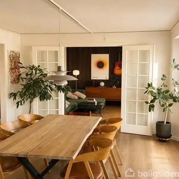 Træspisebord med stole omkring står i forgrunden, og igennem franske døre ses en stue med sofa, vægkunst og guitar. Grønne planter pynter i rummet.