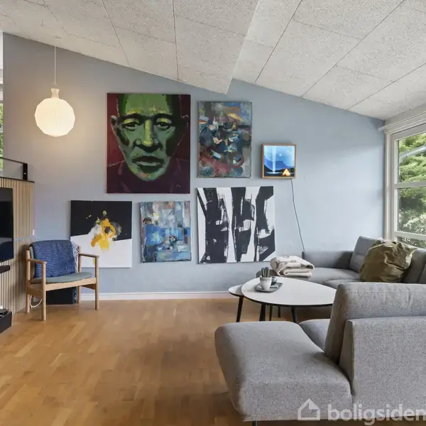 En moderne stue med grå sofa, hængende kunstværker på væggen, fladskærms-tv placeret til venstre og store vinduer, der giver udsigt til en grøn have.