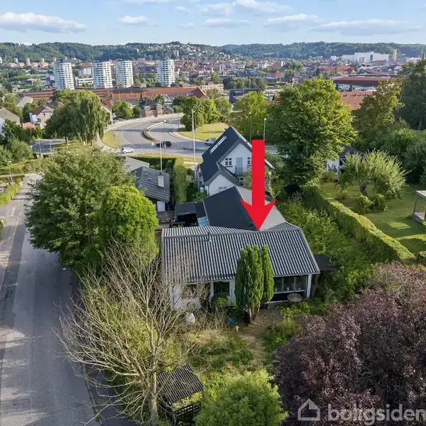 Et hus med en mørk tag, pila peger mod huset; omgivet af træer og en have. Bydelen har flere huse og bygninger i baggrunden med en naturskøn udsigt.