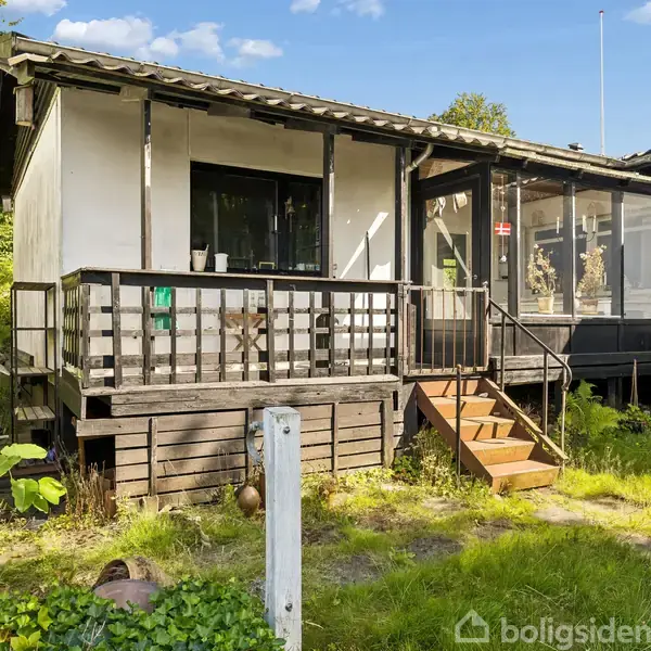 Et lille hvidt hus med en veranda og trappe leder op til en indgang. Det står i en grøn have med græs og træer omkring.