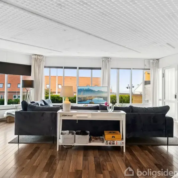 Sofa i stue placeret foran stort vinduesparti, der viser udsigt til rækkehuse. Trægulv strækker sig gennem rummet med en hvid reol og TV i baggrunden.