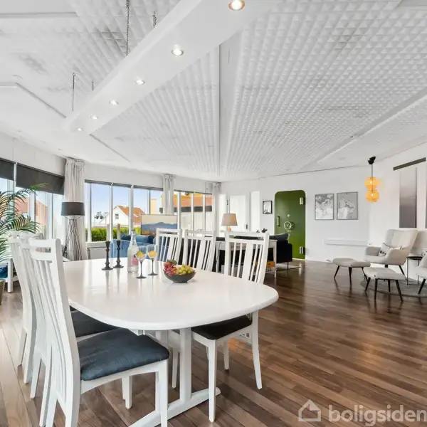 Et lyst spise- og opholdsområde med et stort, ovalt spisebord omringet af hvide stole. Bagved står to komfortable, lyse lænestole. Store vinduer og grønne planter pryder rummet.