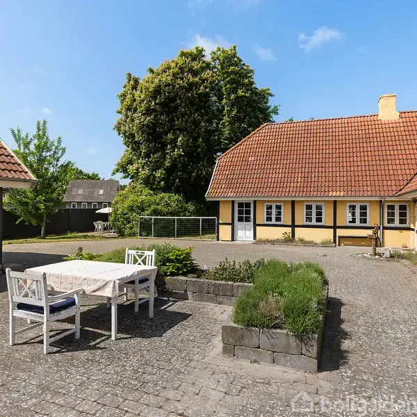 Et gårdmiljø med en udendørs terrasse med bord og stole. Bygninger med gule vægge og røde tegltage omkranser området. Der er træer og buske i baggrunden.