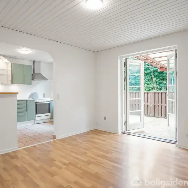 Stue med trægulv åbner sig mod køkken med grønne skabe gennem en bueåbning. Glasdøre fører ud til en terrasse med træhegn, omkranset af grønne planter.