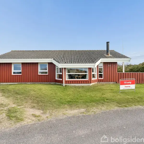 Rødt hus står stille på grøn græsplæne med grussti foran. Skilt viser teksten "Til salg home". Placeringen er naturskøn med buske og sandklitter i baggrunden under en blå himmel.