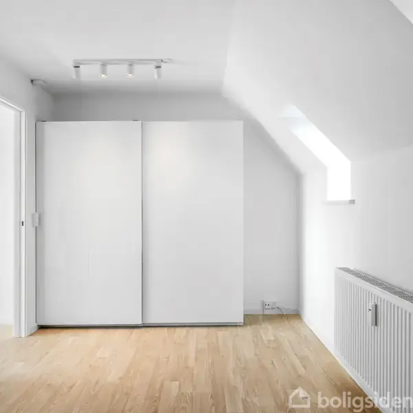 Hvidt skab står mod en væg, under et loftrum med skråvæg og integreret lys. Trægulv dækker rummet, og en radiator er placeret ved et vindue, der giver naturligt lys.
