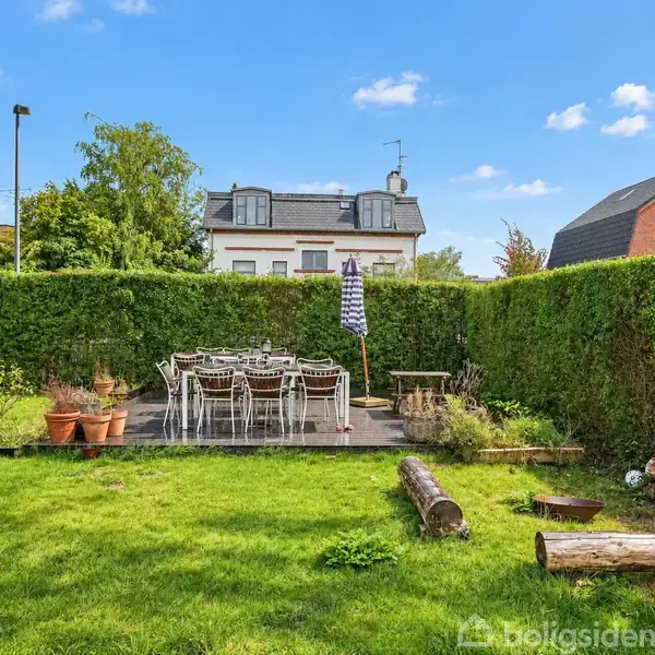 En have med et bord og stole på en terrasse, er omkranset af en grøn hæk. Der er blomsterkrukker og en parasol. I baggrunden ses huse og en blå himmel.