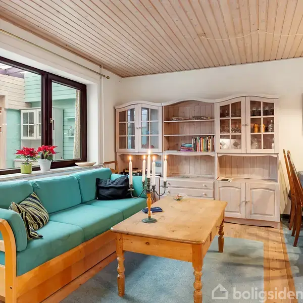 En stue med en turkis sofa står ved et træbord med en kandelaber og stearinlys. Omkring er der en vitrineskab, spisebord med dug og billeder på væggen.