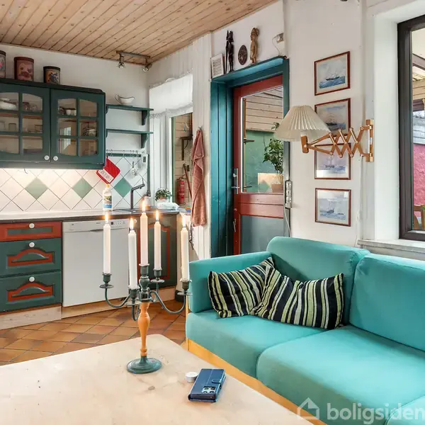 En turkis sofa med stribede puder står i en stue ved siden af et åbent køkken med hvide og grønne skabe. Stearinlys på bordet, dør til højre.