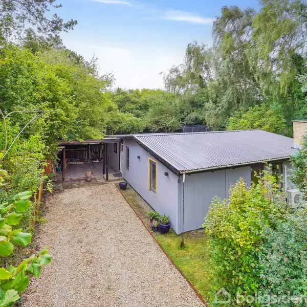 Et enkelt, gråt hus med fladt tag står omgivet af frodig vegetation og træer. En grussti fører op til huset, og bagved er en lille overdækning.