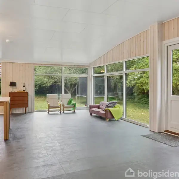 Et lyst rum med spisebord og stole i venstre side, en lænestol til højre, store vinduer og dør, udsigt til grøn have.