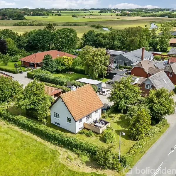 Hus med rødt tag, omgivet af grønne træer og hække, står ved en stille vej i et landsbyområde. Omkringliggende landskab består af marker og spredte bygninger under en skyet himmel.