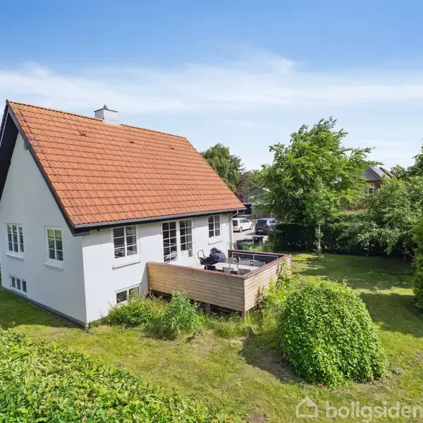 Et hvidt hus med rødt tegltag står i en frodig have. En træterrasse er ved siden af huset. Himlen er klar og blå, omgivet af tæt grøn vegetation.