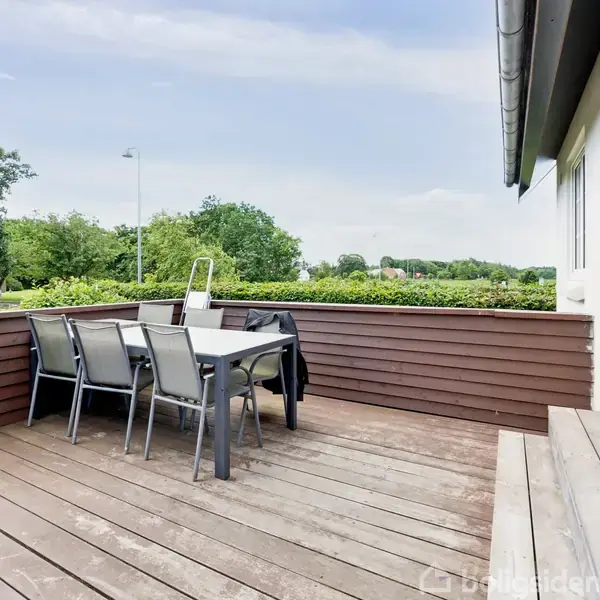 Terrasse med hvidt bord og seks stole i gråt metal står på gulv af træbrædder. Det omkranses af brune træbænke. I baggrunden ses grønne træer og buske.