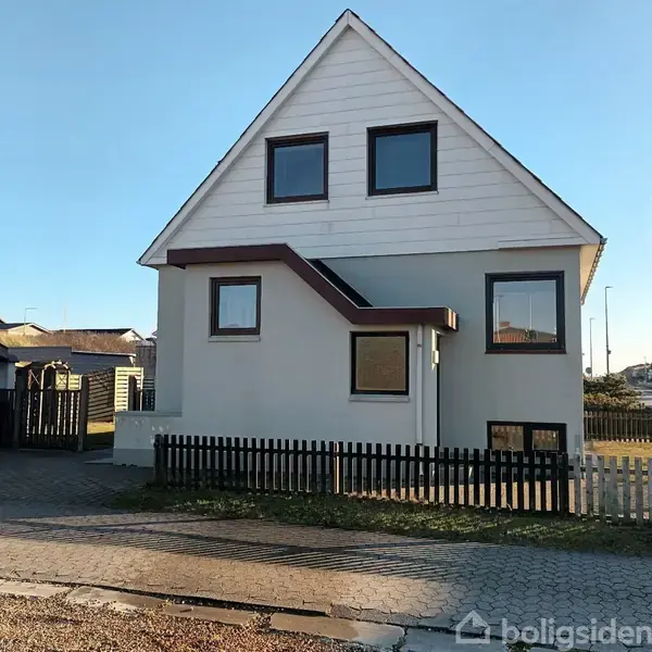 En to-etagers hvid bygning med mørke vinduesrammer står med en skærmtrappe på siden. En sort og hvid stakit omkranser græsplænen. I baggrunden ses flere boligbygninger.