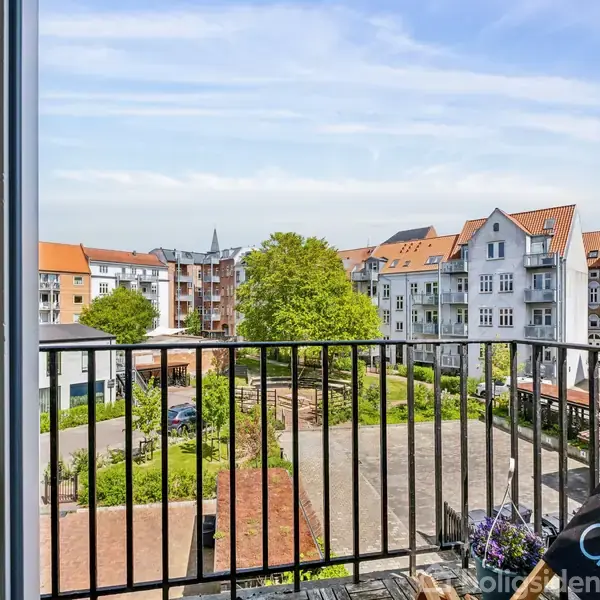 Åben dør fører ud til en lille balkon med udsigt over en gård omgivet af flere etageejendomme med røde tage. Grønne træer og parkerede biler i baggrunden.